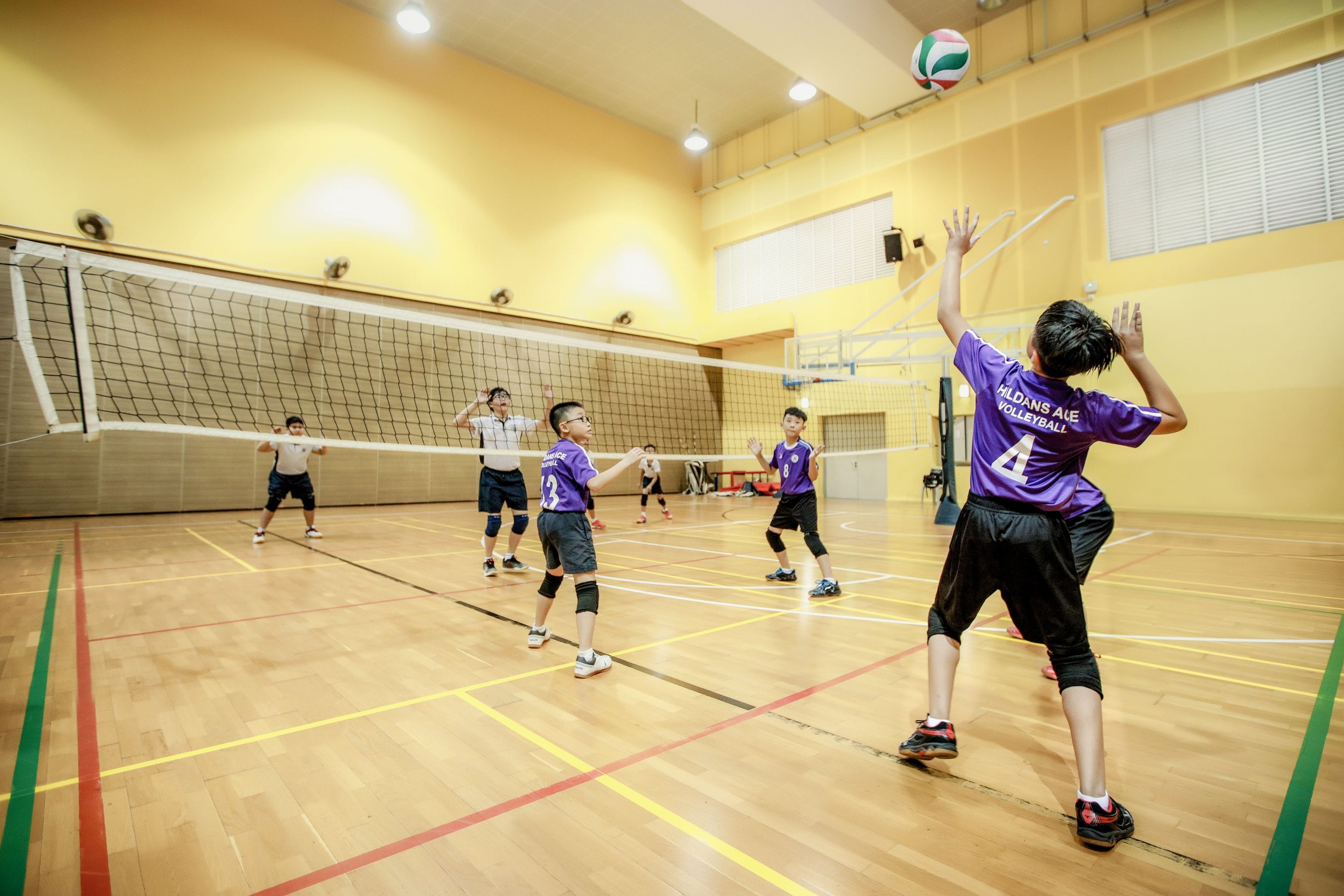 Children in purple volleyball jerseys play indoors, with one player setting the ball above the net.